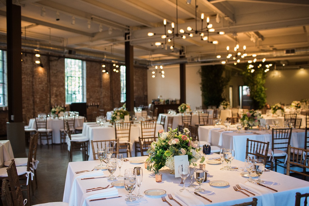 square tables with white table cloths, set for a wedding reception, under industrial style lighting